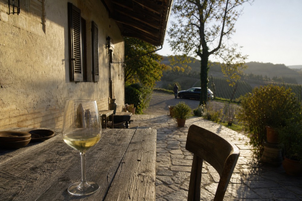 A white wine glass resting on a wooden table outside a stone farmhouse in the Roero hills.