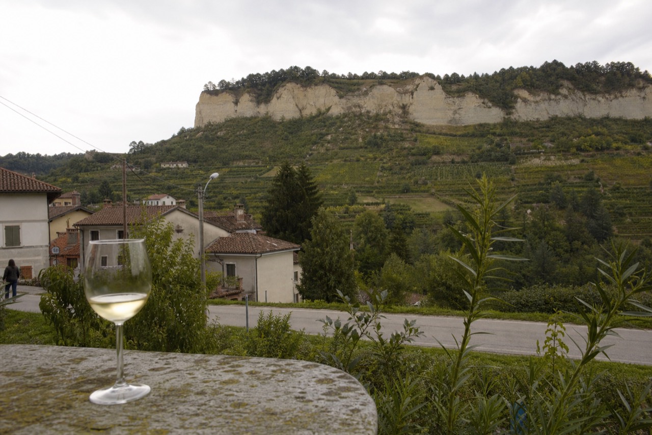 A glass of pale white wine on a stone table beside a vineyard hillside.