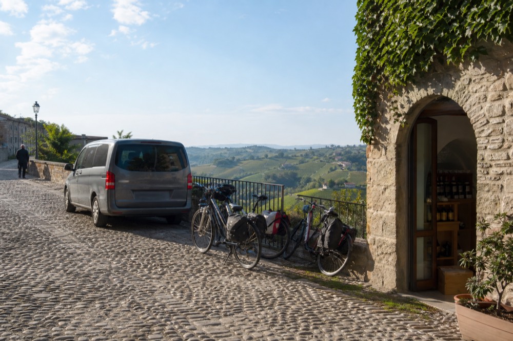 A minivan parked beside a rack of e-bikes in a stone village square above the Barolo hills.