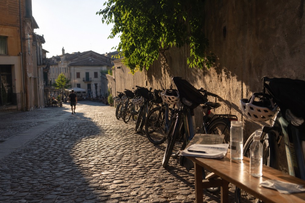 Rental e-bikes propped along a shaded wall in Alba, helmets on the saddles and bar bags open.